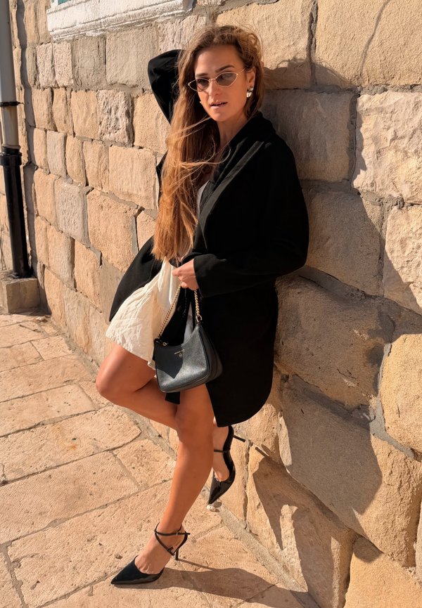 Donna con capelli lunghi che indossa occhiali da sole, cappotto nero, abito bianco e tacchi neri si appoggia a un muro di pietra tenendo una borsa nera al sole.