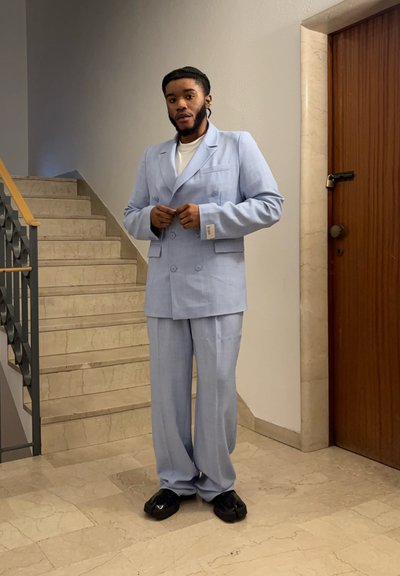 Traje de doble botonadura azul claro con acabado texturizado, pantalones de pierna ancha relajados y zapatos negros. De pie en escaleras de mármol cerca de una puerta de madera.