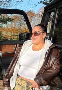 Braune Lederjacke über einem grauen Kapuzenpullover, weißem Oberteil und Tarnhose. Accessoires umfassen große Sonnenbrillen und eine kleine Handtasche. Tür eines schwarzen Fahrzeugs ist geöffnet.