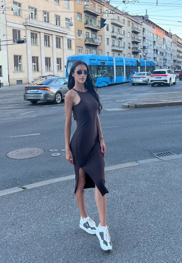 Femme en robe noire et baskets blanches debout dans une rue de la ville avec un tram bleu et des voitures en arrière-plan près des bâtiments urbains.
