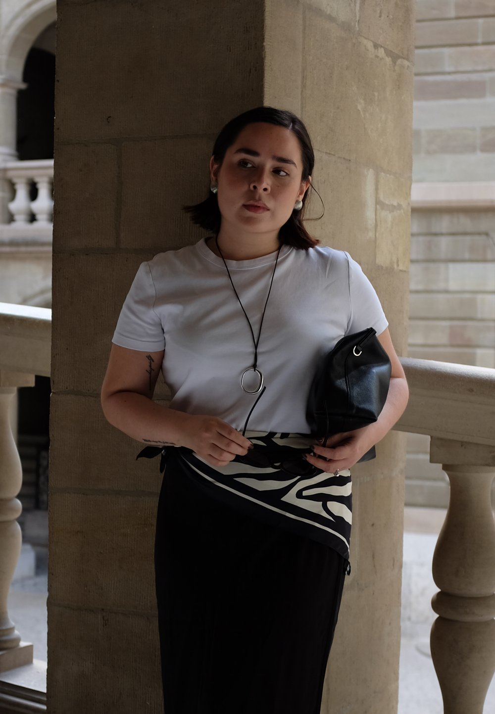 T-shirt bianco a maniche corte, gonna nera con motivo astratto bianco e borsa in pelle nera. La persona è in piedi contro un pilastro di pietra.