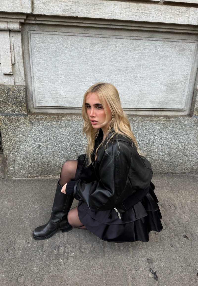 Veste aviateur en cuir noir, jupe noire à volants, bottes noires jusqu'aux genoux. Longs cheveux blonds, assise contre un mur en pierre, en plein air.