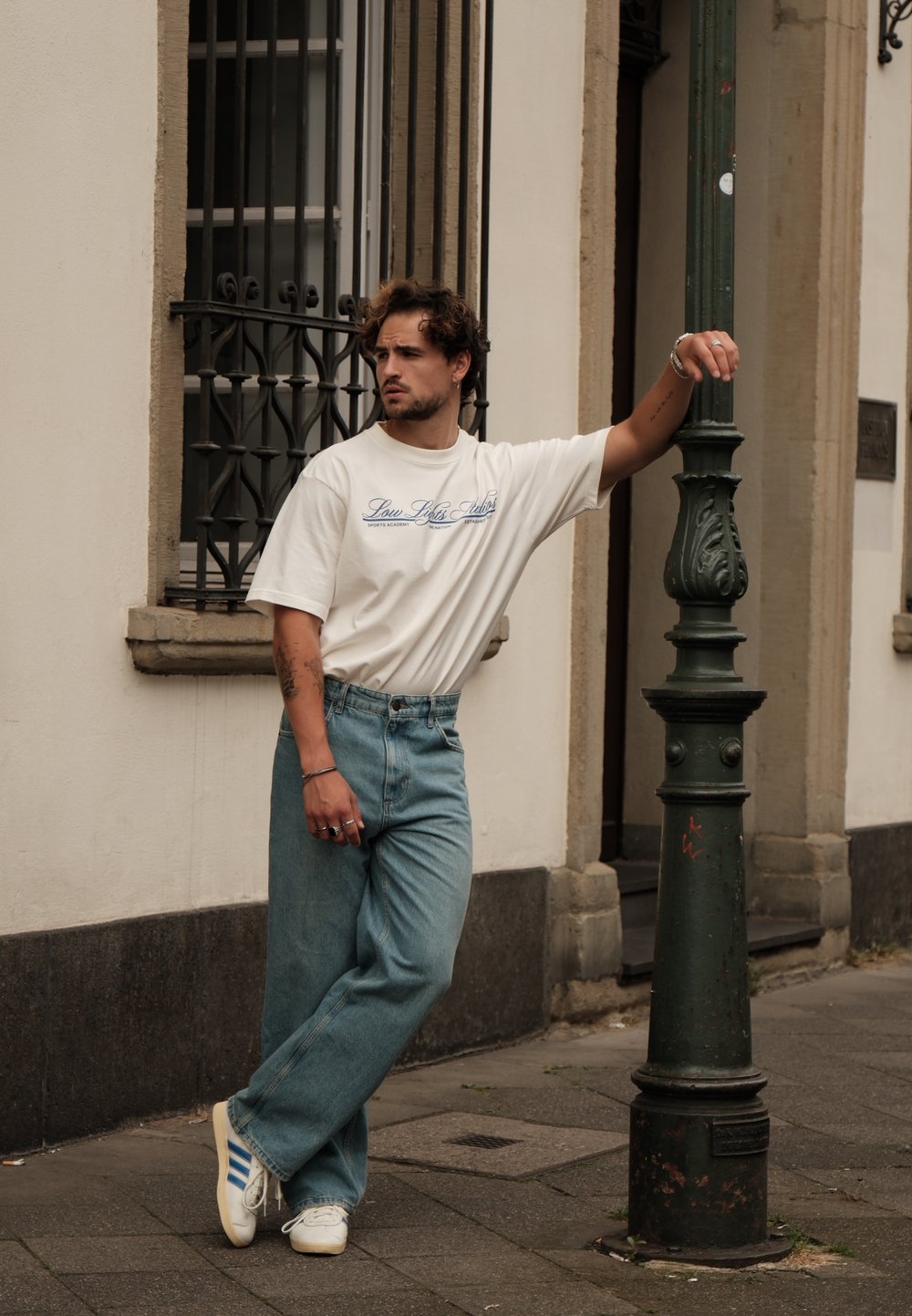 Χαλαρή ενδυμασία με μια κρεμ μπλούζα oversized με μπλε κείμενο, ανοιχτό μπλε φαρδιά τζιν και λευκά αθλητικά παπούτσια με μπλε ρέλι. Στηρίζει σε μια πράσινη κολόνα.