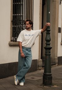 Χαλαρή ενδυμασία με μια κρεμ μπλούζα oversized με μπλε κείμενο, ανοιχτό μπλε φαρδιά τζιν και λευκά αθλητικά παπούτσια με μπλε ρέλι. Στηρίζει σε μια πράσινη κολόνα.