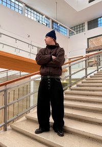 Jeune homme en veste matelassée sombre, pantalon noir ample et bonnet rayé, debout les bras croisés sur un large escalier dans un bâtiment moderne.