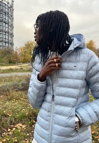 Šviesiai mėlyna pūkinė striukė su horizontaliais siūlėmis, užtrauktuko uždarymu ir gobtuvu, dėvima su balta palaidine. Aksesuarai apima žiedus ir apyrankes.