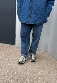 Κουβερτισμένο μπλε μπουφάν, φαρδιά μπλε τζιν παντελόνια και γκρι αθλητικά παπούτσια με λευκές λεπτομέρειες. Στεκόμενος σε μια υφασμάτινη επιφάνεια δίπλα σε έναν τοίχο.