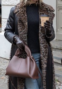 Bolsa de couro castanha com uma textura suave e silhueta única, combinada com um casaco de padrão de leopardo e um suéter preto, segurada por uma mão com luva.