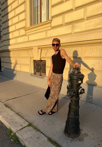 Top sin mangas negro, pantalones con estampado de leopardo, sandalias oscuras; apoyada en una boca de incendio negra contra una pared de piedra texturizada.