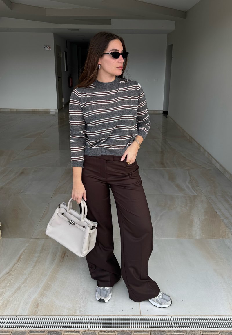Femme portant un pull rayé et un pantalon large marron, tenant un sac à main beige, portant des lunettes de soleil, debout dans un couloir carrelé.