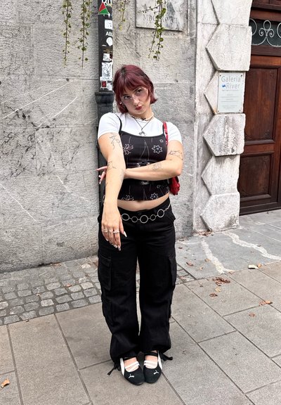 Pantalones de mezclilla negros tipo overall con bordado floral, superpuestos a una camiseta blanca; cinturón de cadena plateada, zapatos de montar con tiras blancas.