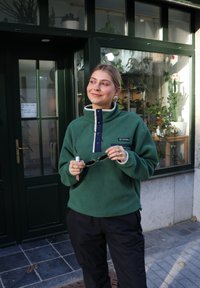Sudadera verde de forro polar con cuello abotonado azul marino y detalles en blanco, combinada con pantalones negros. Persona sostiene gafas de sol frente a una tienda.
