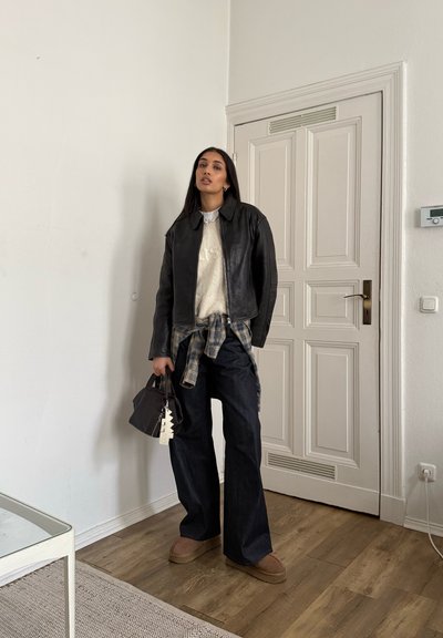Chaqueta de cuero negra sobre un suéter crema, con una camisa azul a cuadros atada a la cintura. Jeans oscuros y holgados, zapatos marrones sin cordones y un bolso pequeño.
