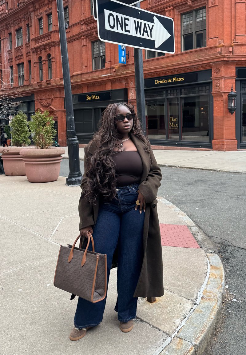 Femme aux longs cheveux, lunettes de soleil, manteau foncé et jeans, tenant un sac à main, debout sur un trottoir en ville sous un panneau de sens unique.