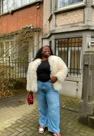 Mujer segura de sí misma que lleva un abrigo blanco peludo, camiseta negra, vaqueros azules y zapatos rojos, sosteniendo un bolso rojo, de pie cerca de un edificio de ladrillo y piedra.