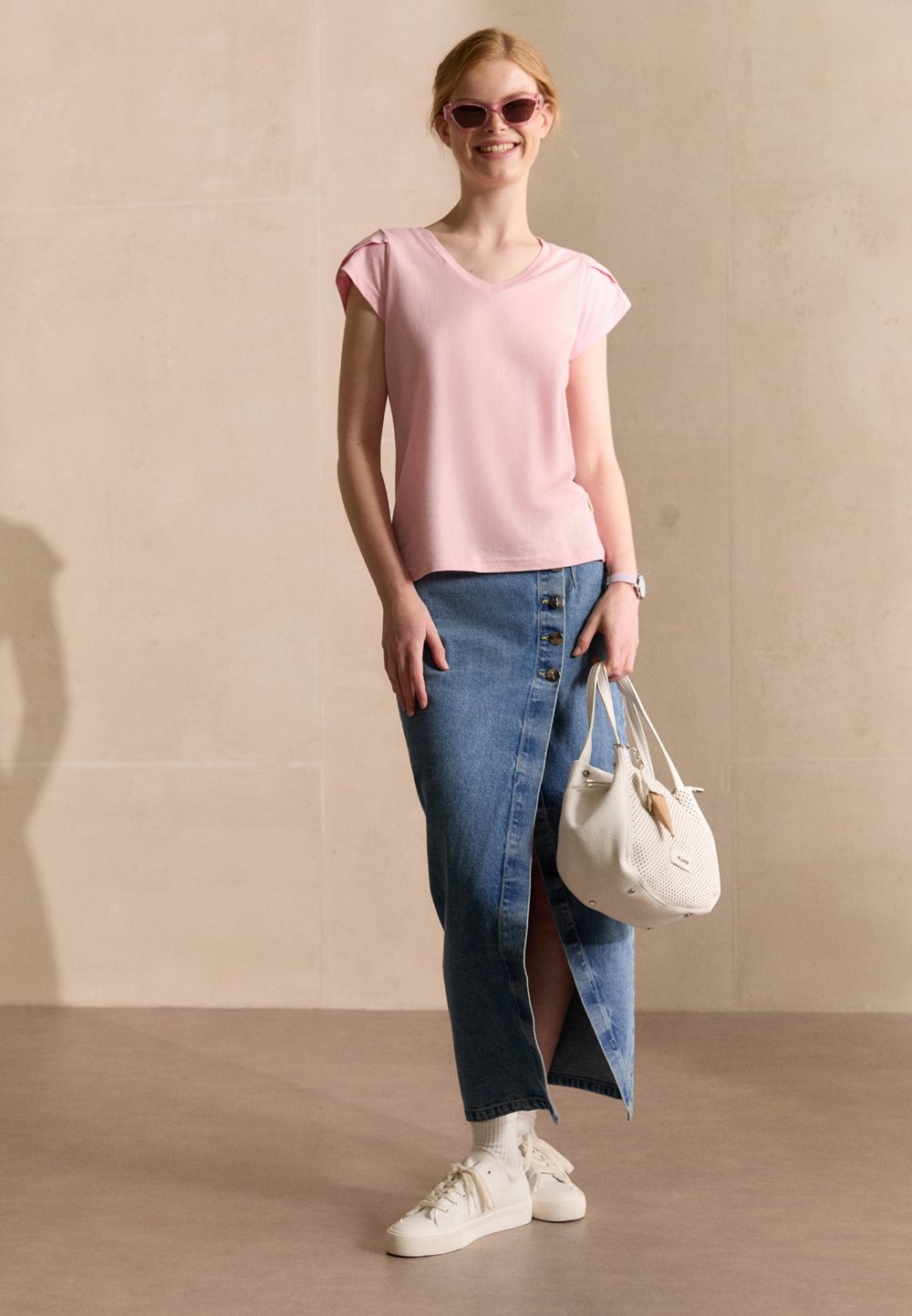 Top rosa a maniche corte con scollo a V, gonna lunga in denim con bottoni frontali e spacco, borsa bianca e sneakers bianche con design testurizzato.