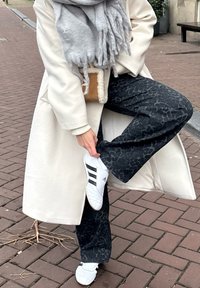 White long coat over a grey scarf, black trousers with a mottled pattern, and white trainers with black stripes, standing on brick pavement.