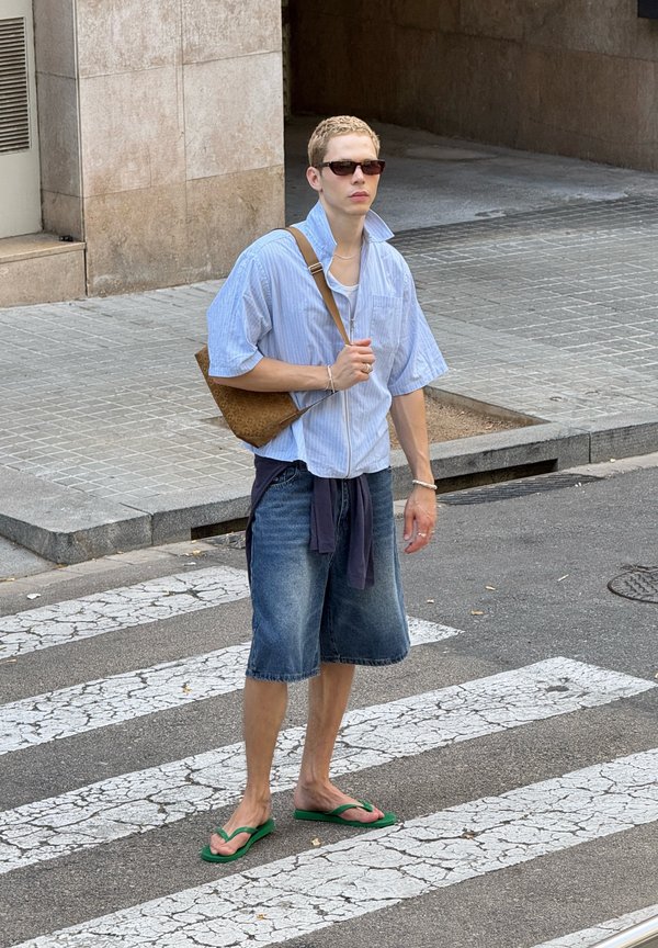 Giovane persona con occhiali da sole, camicia blu, pantaloncini di jeans e infradito verdi, in piedi su un attraversamento pedonale in città con una borsa a tracolla marrone.