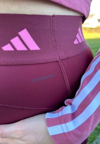 Leggings atletici color bordeaux con logo Adidas rosa, caratterizzati da una texture liscia e un'etichetta "AEROREADY" sulla vita. Vista ravvicinata.