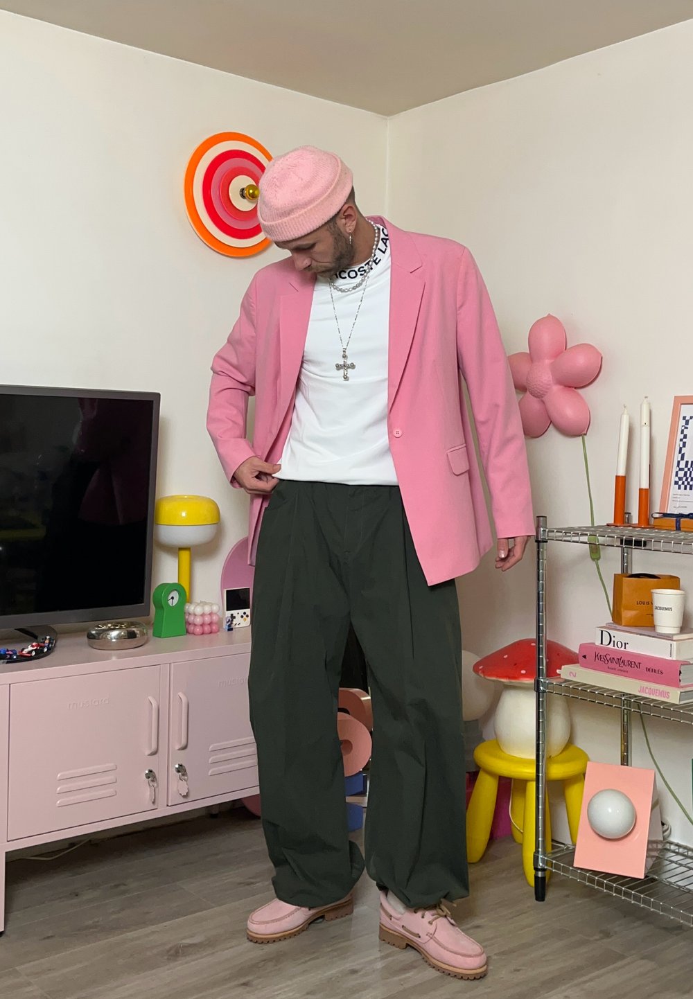 Rosa kavaj över vit skjorta, gröna vida byxor, ljusrosa skor och en rosa beanie. Enkel korshalsband och en färgglad inredning.