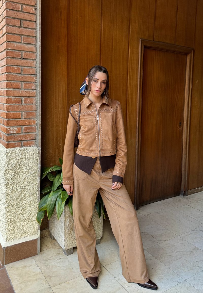 Braune Wildlederjacke mit Nietenakzenten, kombiniert mit passenden braunen Hose mit weitem Bein. Strukturierter, lockerer Schnitt. Dunkelbraune Bündchen und Saumdetail.