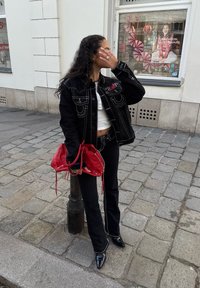 Jaqueta de ganga preta com costura em contraste, top curto branco, calças pretas largas e sapatos de salto pretos brilhantes; segurando uma bolsa vermelha com cordão.