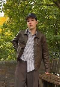 Jaunas vyras rudos odinės striukės ir dryžuoto marškinėliais stovi lauke prie medinio suolo ir žaliuojančių medžių.