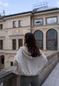 Lekki, kremowy hoodie z kapturem ze sznurkiem, opadającymi ramionami i elastycznym dołem, w połączeniu z dopasowanymi, zielonymi legginsami, stojący na zewnątrz.