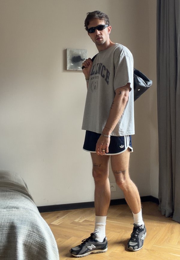 Man wearing sunglasses, gray t-shirt, black shorts, and sneakers, carrying a black bag over his shoulder standing indoors near a bed and curtain.