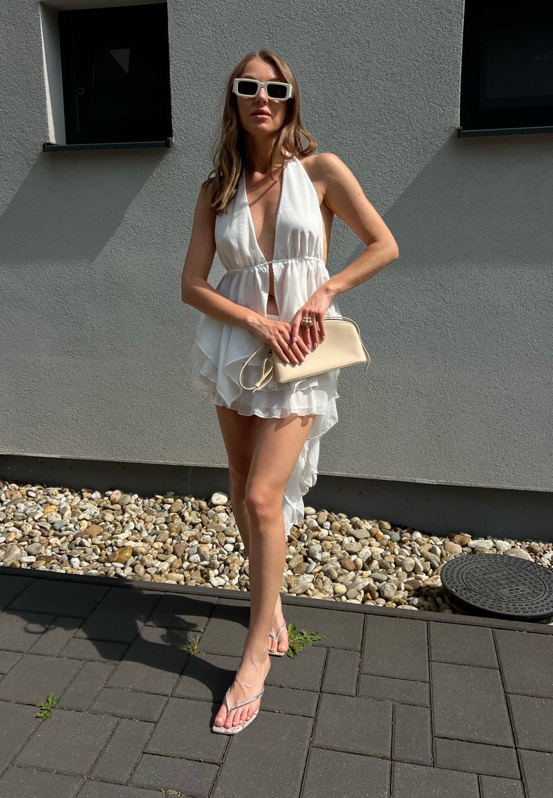 Femme portant une robe dos nu blanche, des lunettes de soleil blanches et des sandales se tient sur la pierre et le trottoir en tenant une pochette beige contre un mur gris.