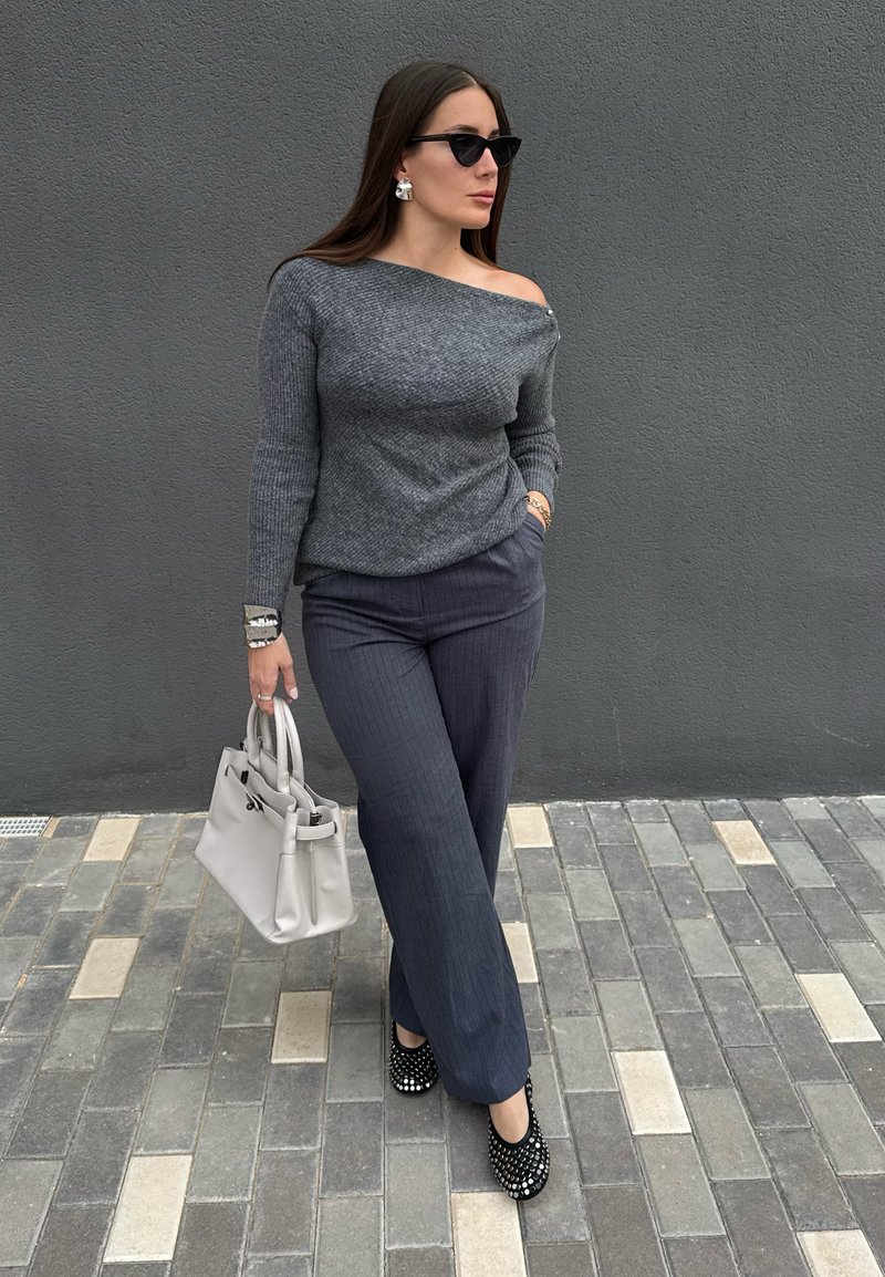 Pull gris côtelé à épaules dénudées, pantalon tailleur foncé à fines rayures, talons noirs cloutés et sac à main gris clair. Textures lisses, design moderne.