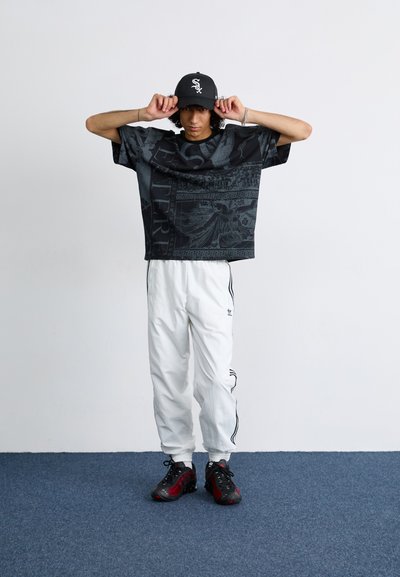 Camiseta gráfica negra de gran tamaño con patrones claros, pantalones blancos ajustados con rayas negras y zapatillas deportivas negro-rojas.