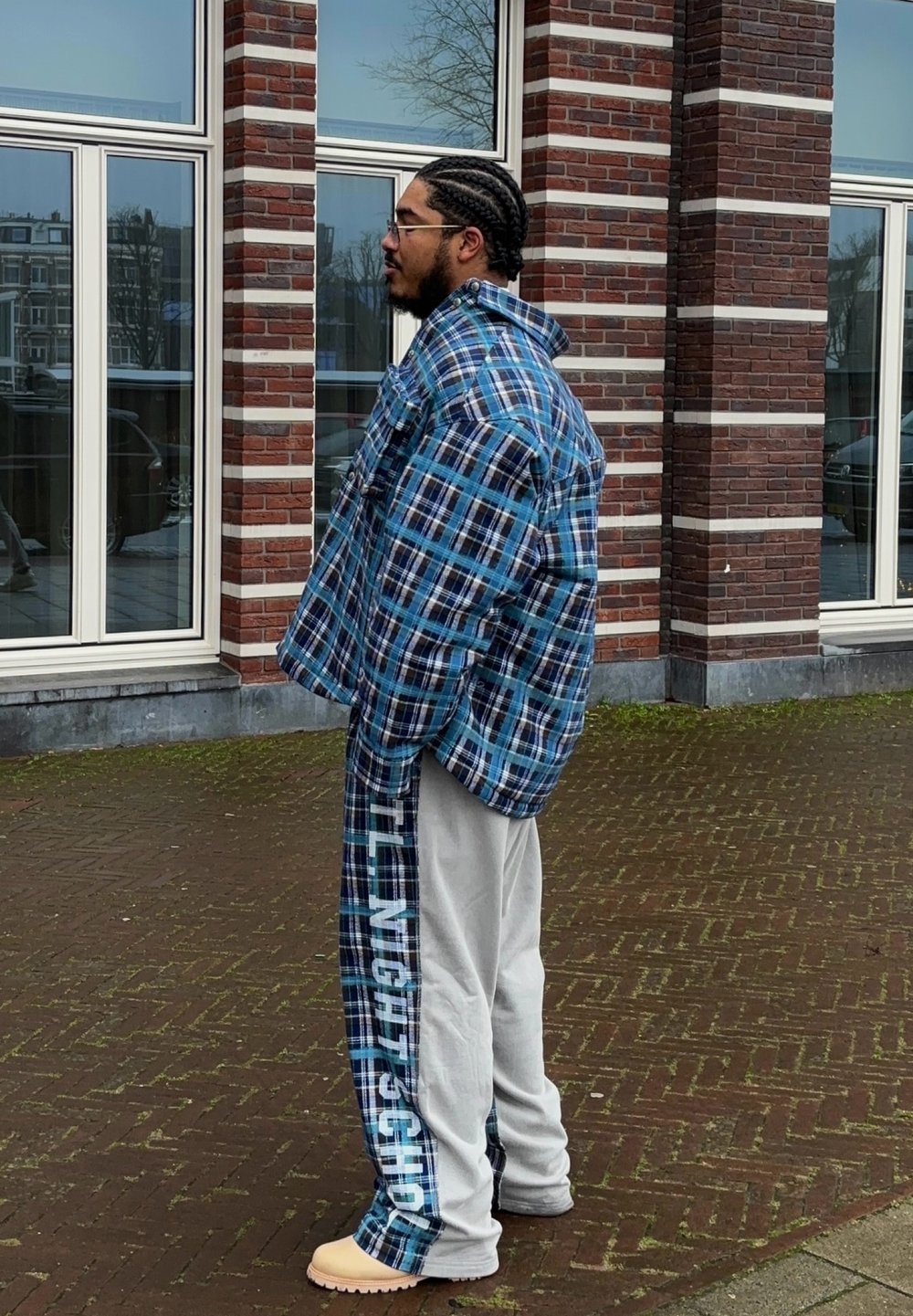 Casaco à prova de vento azul de padrão xadrez oversized sobre calças de moletom cinzas com texto branco. Calçado bege com sola texturizada. Parede de tijolo e janelas de vidro ao fundo.
