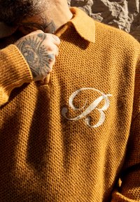 Pull en tricot couleur moutarde avec un motif texturé et un col à revers. Présente une lettre "B" blanche brodée sur la poitrine.
