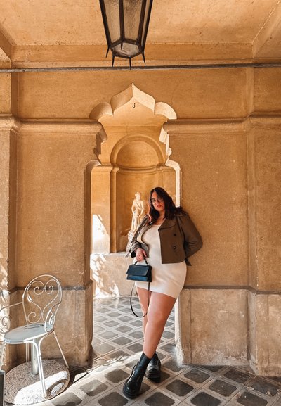 Chaqueta marrón de doble botonadura, vestido blanco de canalé, bolso mini negro, botas negras sólidas, paredes de piedra beige claro, arco decorativo, suelo con patrón.