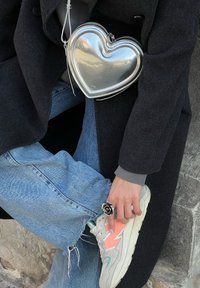 Silverfärgad handväska i form av ett hjärta med en glänsande yta, dragkedjor och ett tunt axelremsband. Bärs tillsammans med ljusblå jeans och grå sneakers.