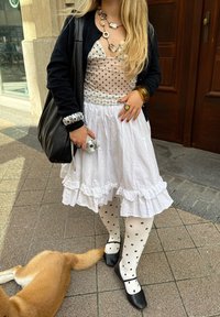 Gonna bianca a balze, collant neri a pois, ballerine nere, top trasparente a pois, collana a strati, cardigan nero, mentre tiene una macchina fotografica.