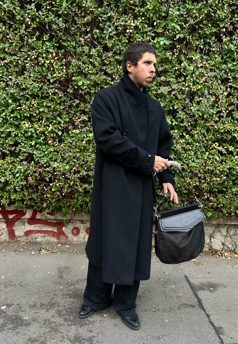Man in long black coat and black pants holding black handbag, standing on sidewalk in front of dense green hedge with red graffiti on low wall.