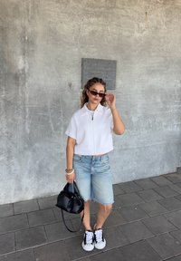 Giacca corta bianca con zip, pantaloni corti di jeans, borsa nera e occhiali da sole neri. Indossa sneakers bianche e nere. Sfondo grigio strutturato.