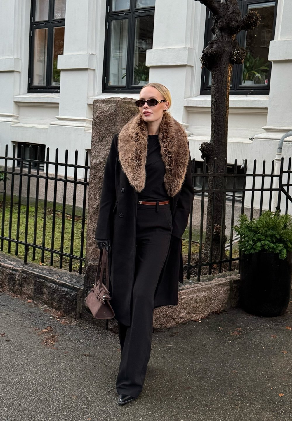 Manteau noir sur mesure avec un col en fausse fourrure, porté par-dessus un haut noir et un pantalon sur mesure. Accessoirisé avec des gants noirs et un sac à main marron.