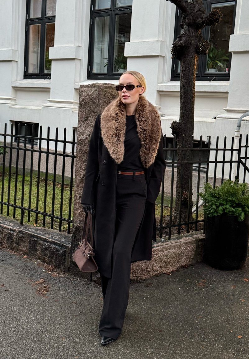 Manteau noir sur mesure avec un col en fausse fourrure, porté par-dessus un haut noir et un pantalon sur mesure. Accessoirisé avec des gants noirs et un sac à main marron.