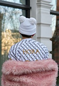 Gorro de malha branco com dois pompons, acompanhado de uma camisa de manga curta com padrão que apresenta o texto dourado "HORA" e uma jaqueta rosa fofa.