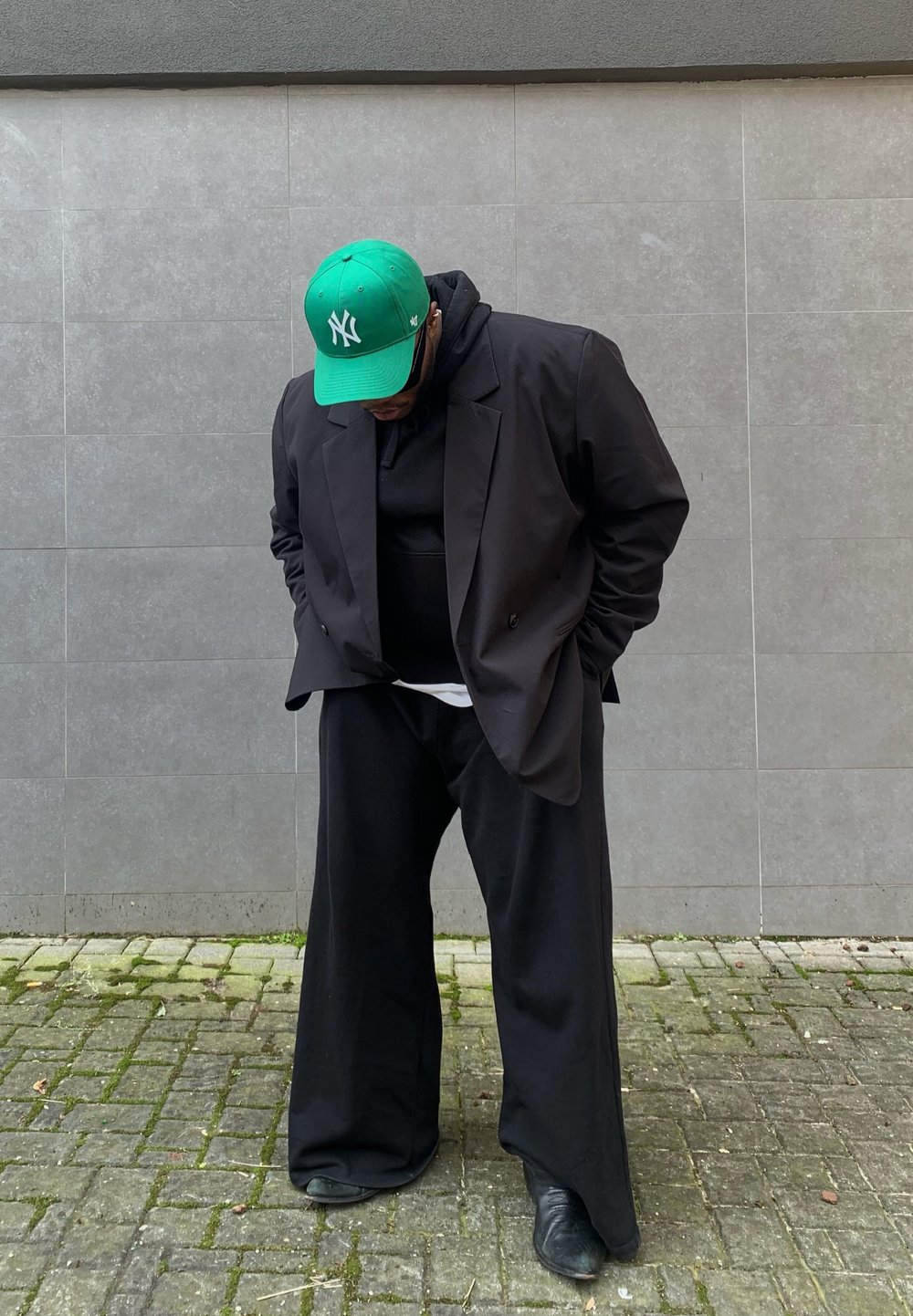 Czarna oversize'owa marynarka, czarna hoodie pod spodem, szerokie czarne spodnie oraz zielona czapka New York Yankees, do tego błyszczące czarne buty. Szary tle.