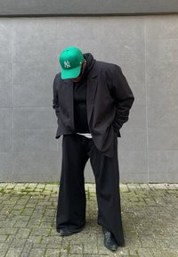 Czarna oversize'owa marynarka, czarna hoodie pod spodem, szerokie czarne spodnie oraz zielona czapka New York Yankees, do tego błyszczące czarne buty. Szary tle.