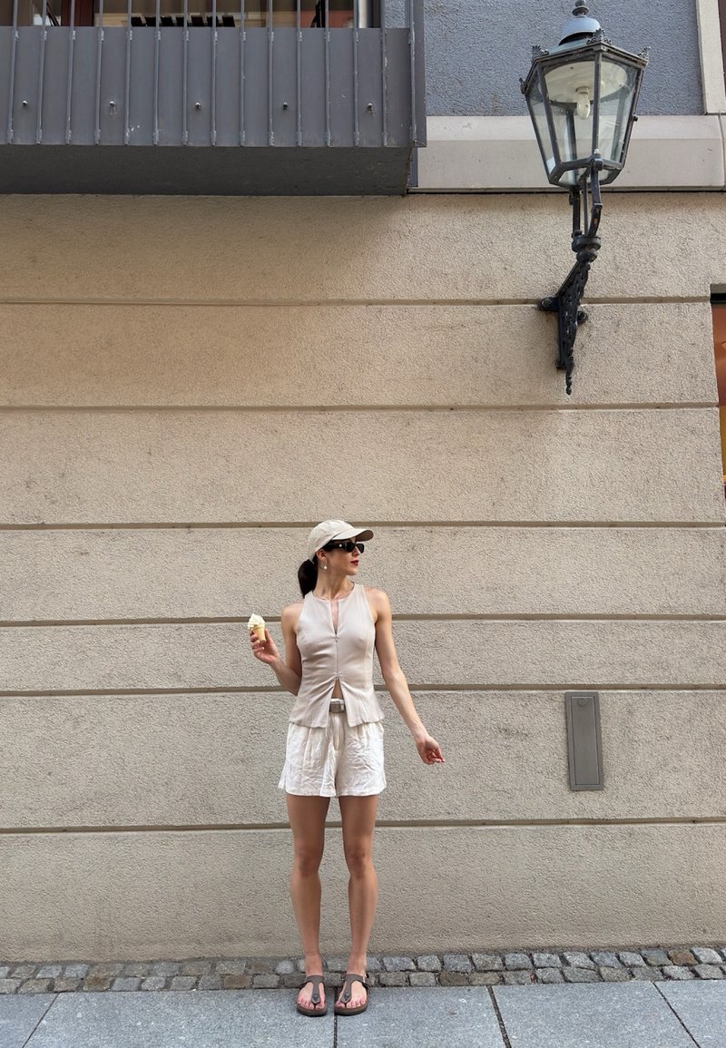 Woman in beige top, white shorts, cap, and sandals holding an ice cream cone standing against a beige stone wall under a street lamp.
