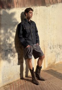 Tume denim jakk koos kontrastsete kollaste õmblustega, hallid cargo lühikesed püksid ja pruunid cowboy saapad. Seisab reljeefse seina vastas.