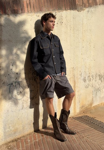 Chaqueta de mezclilla oscura con costuras amarillas en contraste, pantalones cortos de carga grises y botas de vaquero marrones. De pie contra una pared texturizada.