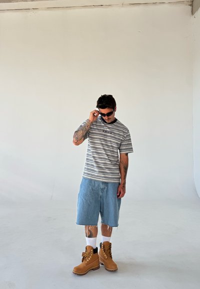 Camiseta de rayas de manga corta, pantalones cortos de denim azul claro, calcetines blancos hasta el tobillo, botas de trabajo marrones con cordones amarillos, sobre un fondo blanco minimalista.