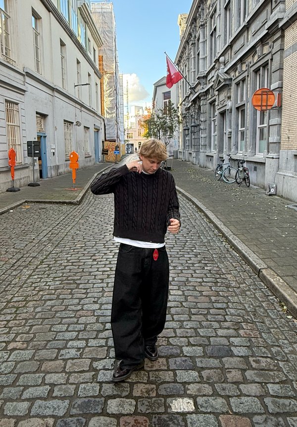 Schwarzer Kabelstrickpullover, weißes T-Shirt darunter, lockere schwarze Hose. Steht auf einer Pflasterstraße mit Gebäuden und Bäumen im Hintergrund.