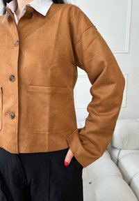 Veste en daim marron avec ouverture boutonnée, col et une poche poitrine unique. Texture lisse, coupe décontractée et manches longues.